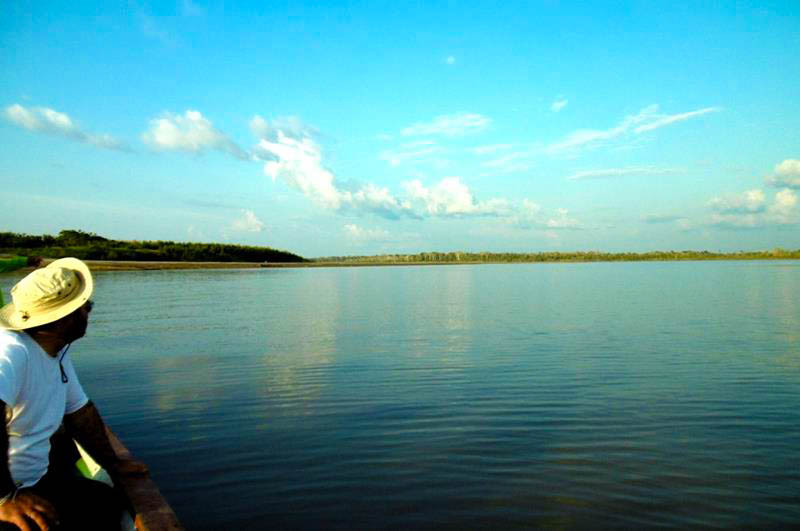 Person sitting in a boat on a calm Amazon river, looking toward the horizon under a clear blue sky with scattered clouds