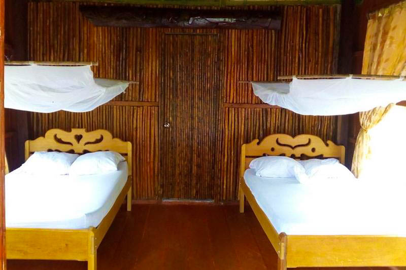 Room at Antares Lodge with two wooden beds, white bedding, mosquito nets, bamboo walls, wooden floor, and a window with light-colored curtains